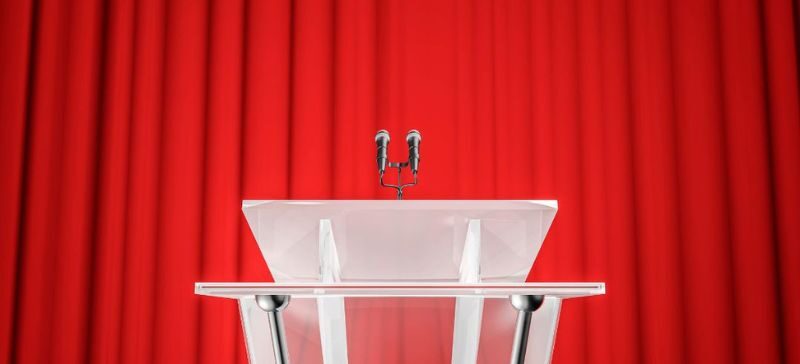 white podium with 2 microphones sits in front of a bright red curtain