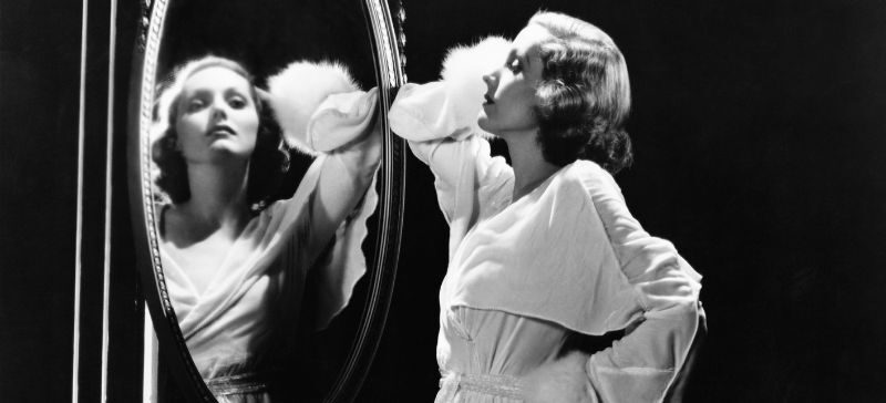 vintage black and white photo where a 1920's woman in a white rode looks at herself in a mirror