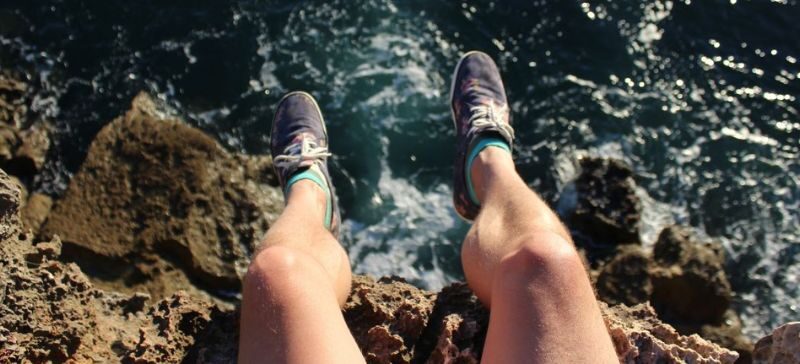 in a risky move person dangles their feet over the edge of a cliff that looks into ocean