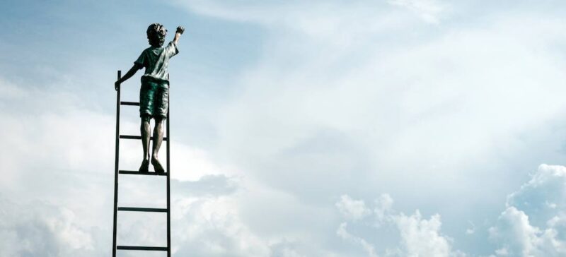 statue of a boy on a ladder reaching for the clouds