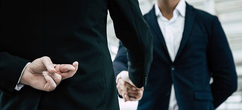 Two men in suits, shaking hands, while one is crossing his fingers behind his back, indicating duplicity